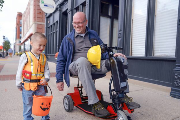 An older man is riding his mobility scooter along the street. Walking alongside is a child playing dress-up as a builder.
