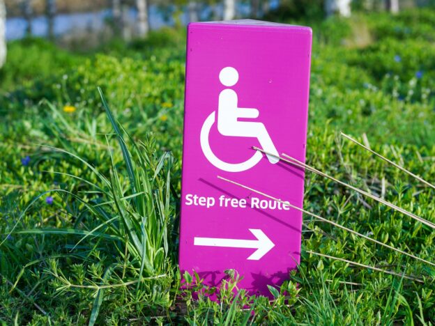 A sign, with a pink background and white foreground, shows a step-free access route and a wheelchair symbol. The sign is positioned on a green lawn.